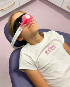 a child patient laying back in a dental chair wearing a pink happy gas sedation mask for calm visits to the dentist 