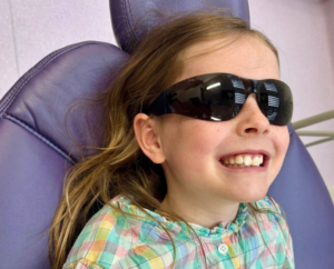 a young girl sitting in a purple dental chair wearing protective glasses smiling at the camera to promote happy and calming dental visits in london