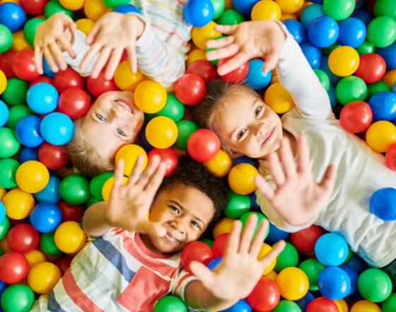 a photo of three young children laying in a colourful ball pitt looking at the camera smiling