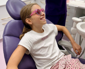 an image of a young girl sitting in a purple dental chair smiling whilst wearing fun pink protective glasses
