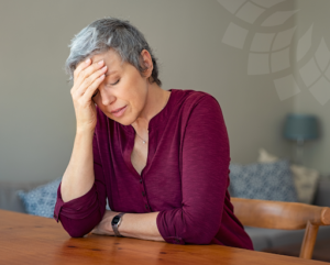 a woman sitting at a table wearing a maroon shirt with her head in her hand to depict the feeling of stress